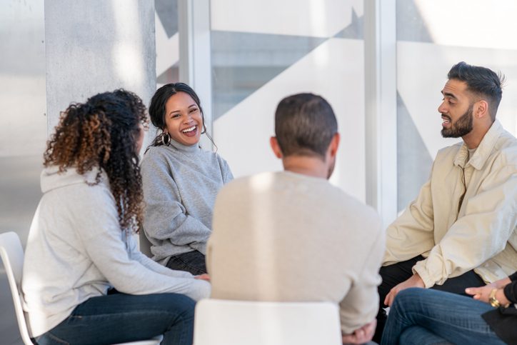 A small group diverse adults sit together as they participate in a group therapy session.  They are each dressed casually as they go around the circle sharing their struggles and supporting one another.