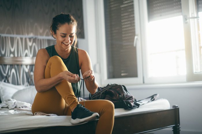 woman in athletic wear tying her shoes while sitting at the edge of the bed. Getting ready to work out.