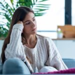 Young woman sitting on a couch indoors, leaning her head on her hand and looking pensive or sad, wearing a white cable-knit sweater.