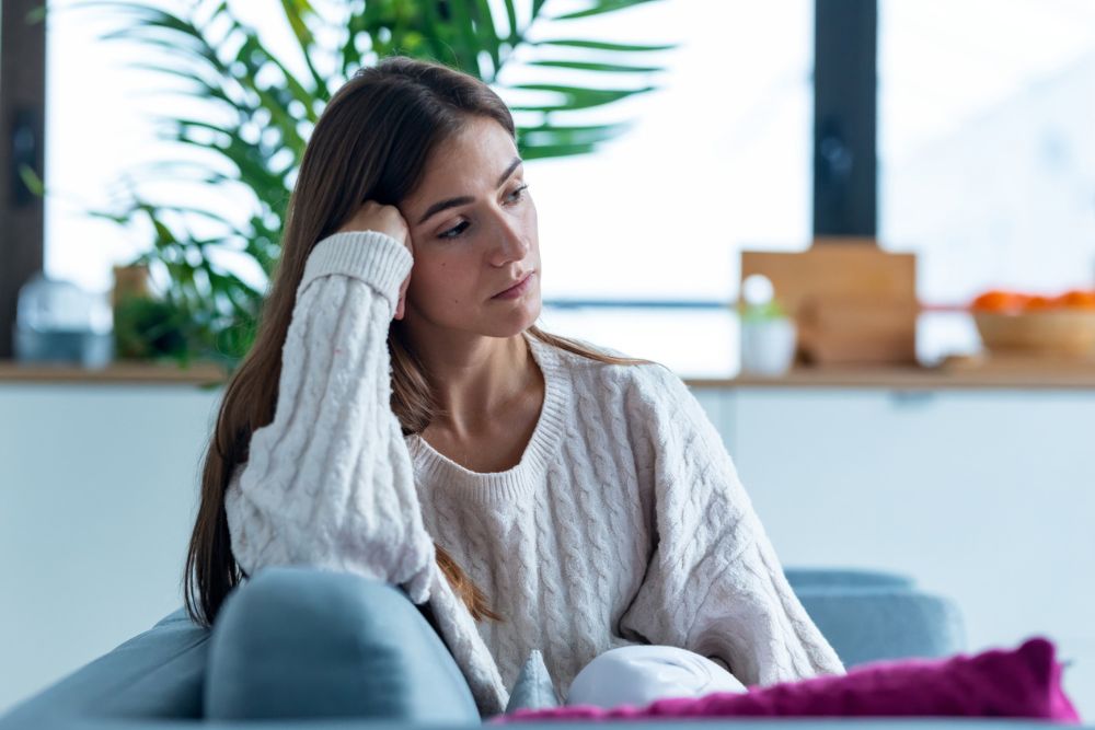 Young woman sitting on a couch indoors, leaning her head on her hand and looking pensive or sad, wearing a white cable-knit sweater.