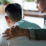 Man offering support by placing a comforting hand on another man's back, symbolizing empathy, compassion, and emotional support during a difficult time.
