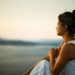 Woman sitting peacefully on a cliffside at sunset, gazing over calm water with a thoughtful expression,