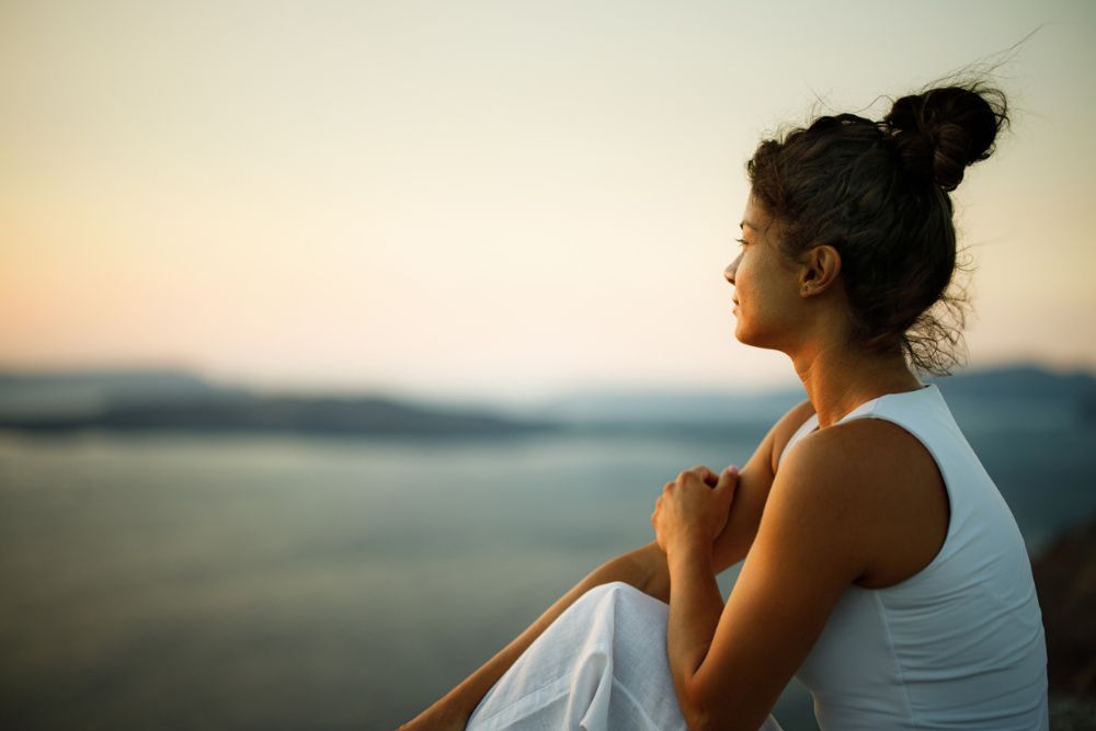 Woman sitting peacefully on a cliffside at sunset, gazing over calm water with a thoughtful expression,