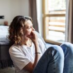 A woman sitting on the floor next to a bed, looking out the window with a pensive expression, suggesting emotional distress or deep thought.