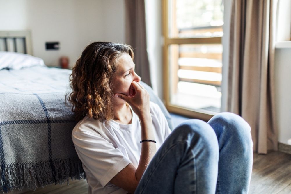 A woman sitting on the floor next to a bed, looking out the window with a pensive expression, suggesting emotional distress or deep thought.