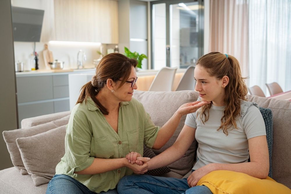 A caring adult woman sits on a couch holding a teenage girl’s hand and gently touching her chin, offering comfort. The teen looks emotional. They are in a bright, modern living room and kitchen space.