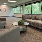A modern outpatient waiting area with gray sofas, armchairs, a round coffee table with a plant, large windows overlooking trees, and abstract wall art.
