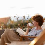 oman sitting on a wooden deck chair, wrapped in a blanket, drinking from a white mug and reading a book outdoors during sunset, with soft sunlight and plants in the background.