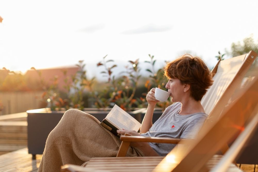 oman sitting on a wooden deck chair, wrapped in a blanket, drinking from a white mug and reading a book outdoors during sunset, with soft sunlight and plants in the background.