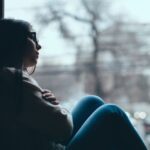 Teen girl sitting by a window with arms wrapped around her knees, looking out thoughtfully, suggesting sadness or depression.