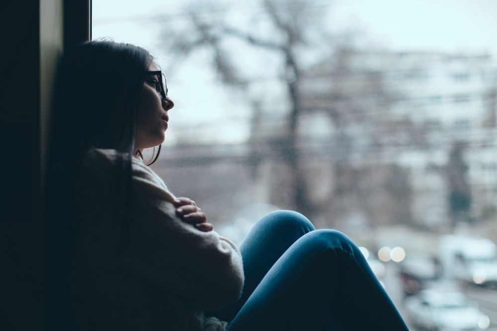 Teen girl sitting by a window with arms wrapped around her knees, looking out thoughtfully, suggesting sadness or depression.