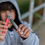 Teenager in a hoodie holding two vape pens outdoors, examining them closely with a blurred background.