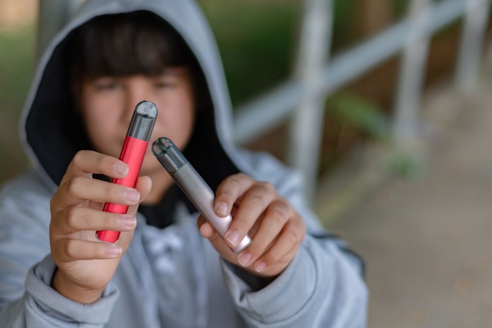 Teenager in a hoodie holding two vape pens outdoors, examining them closely with a blurred background.