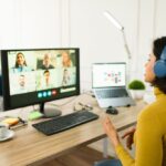 A woman wearing headphones sits at a desk while participating in a video call on her computer, with multiple colleagues visible on the screen. A laptop, coffee cup, and office supplies are on the desk in a bright home workspace.