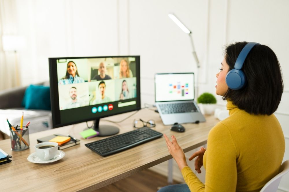 A woman wearing headphones sits at a desk while participating in a video call on her computer, with multiple colleagues visible on the screen. A laptop, coffee cup, and office supplies are on the desk in a bright home workspace.
