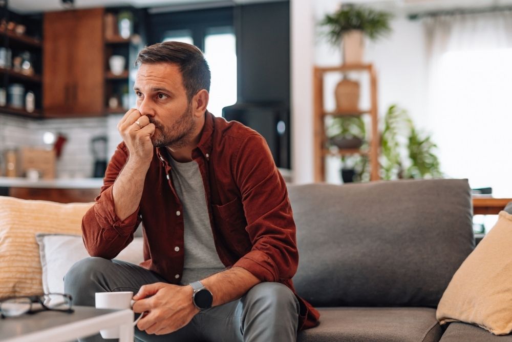 A man sits on a couch holding a mug, resting his hand near his mouth with a worried expression in a quiet, home setting.