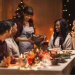 A woman serves a holiday meal to a smiling group of friends seated around a decorated table with warm lights and festive décor