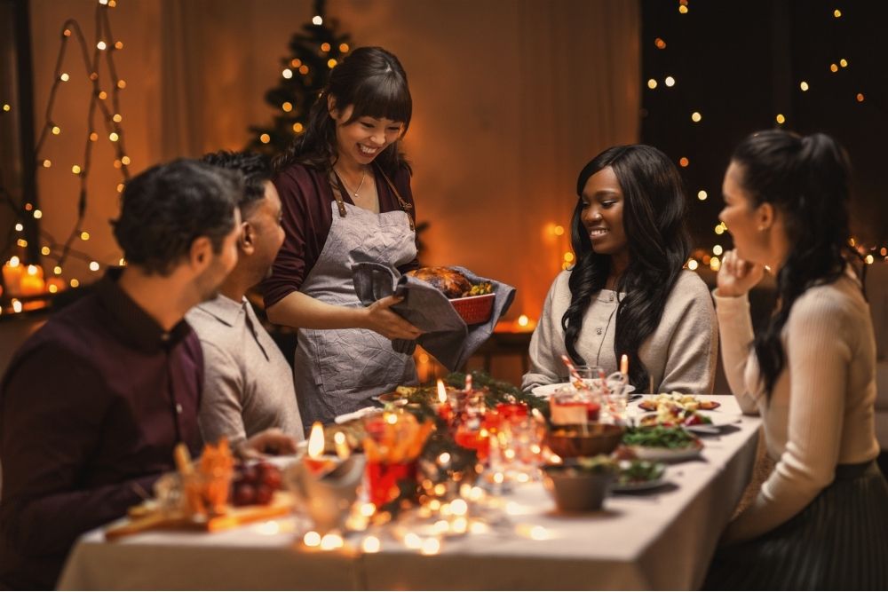 A woman serves a holiday meal to a smiling group of friends seated around a decorated table with warm lights and festive décor