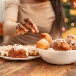 A person reaching for sugary holiday pastries, with a bowl of powdered donuts and waffles on a table.