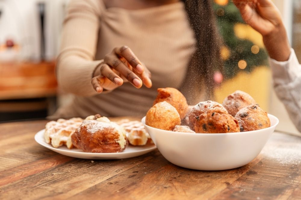 A person reaching for sugary holiday pastries, with a bowl of powdered donuts and waffles on a table.
