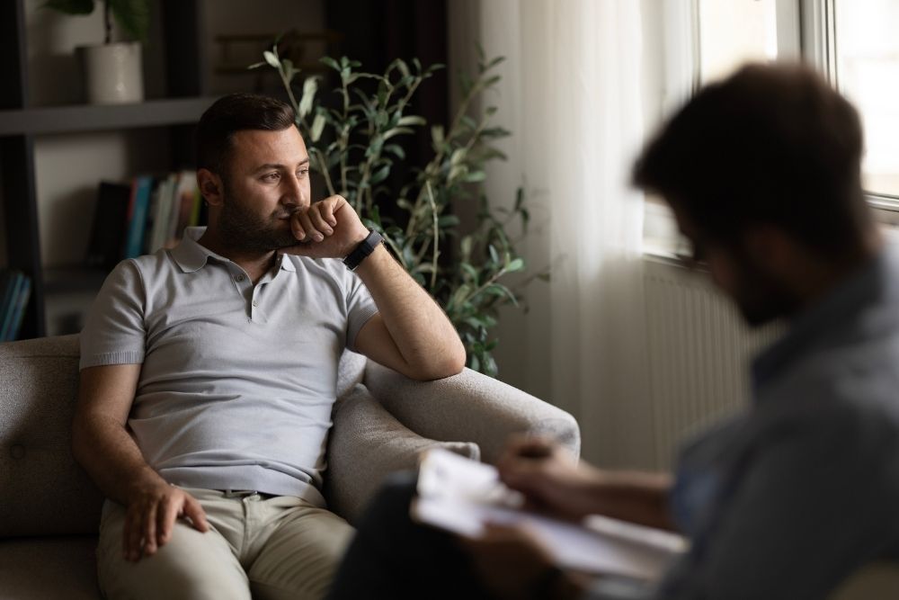 Miramount - Genetics and Dual Mental Health Diagnoses. A young man sits in a therapy session in a private office. He looks out the window in a daze while a therapist writes notes.