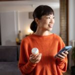 Person in an orange sweater holding a small bottle and a smartphone, standing in a well-lit room with a gray sofa and a plant. The person holds a depiction of AUD medication,
