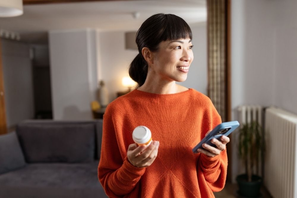 Person in an orange sweater holding a small bottle and a smartphone, standing in a well-lit room with a gray sofa and a plant. The person holds a depiction of AUD medication,